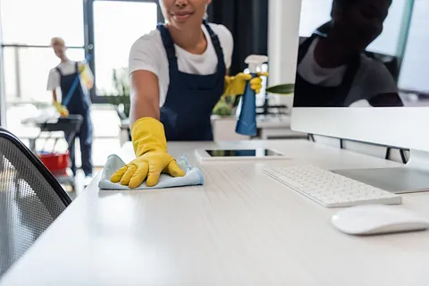 Vista recortada de una mujer birracial con guantes de goma limpiando el escritorio de la oficina cerca de gadgets y un colega en un fondo borroso